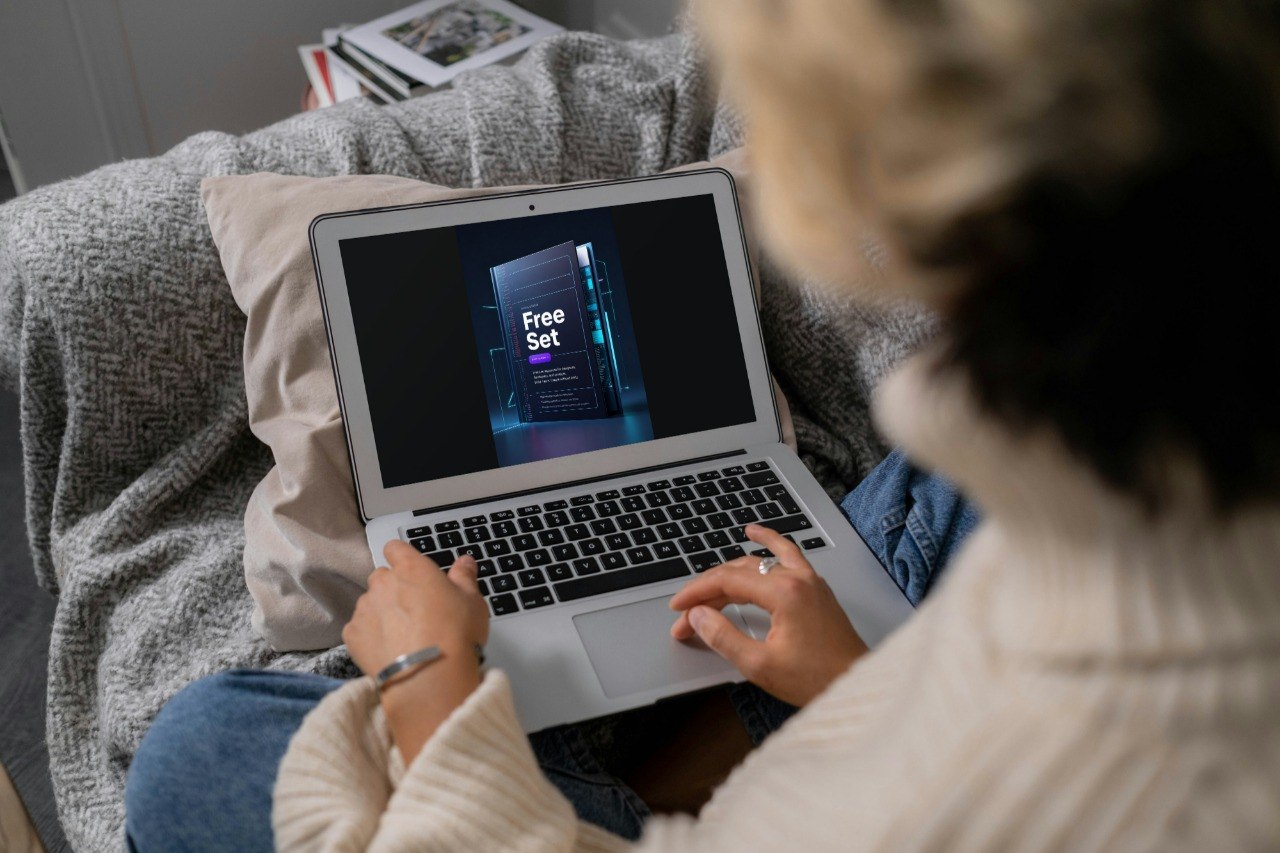 A person sitting on a couch uses a silver laptop showing a digital course cover. The person is wearing a cream ribbed sweater and blue jeans, with a gray textured blanket and pillows around them.