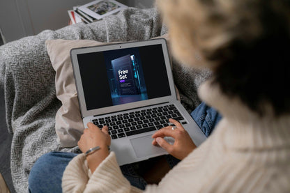 A person sitting on a couch uses a silver laptop showing a digital course cover. The person is wearing a cream ribbed sweater and blue jeans, with a gray textured blanket and pillows around them.