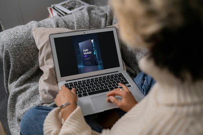 A person sitting on a couch uses a silver laptop showing a digital course cover. The person is wearing a cream ribbed sweater and blue jeans, with a gray textured blanket and pillows around them.